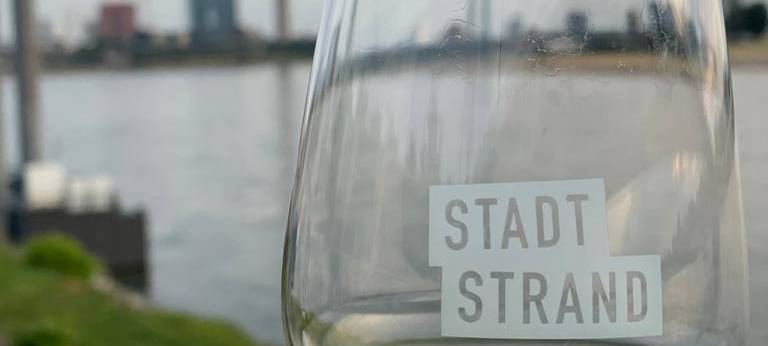 Ein Weinglas mit Weißwein und der Aufschrift "Stadtstrand", im Hintergrund ist die Düsseldorfer Skyline zu sehen