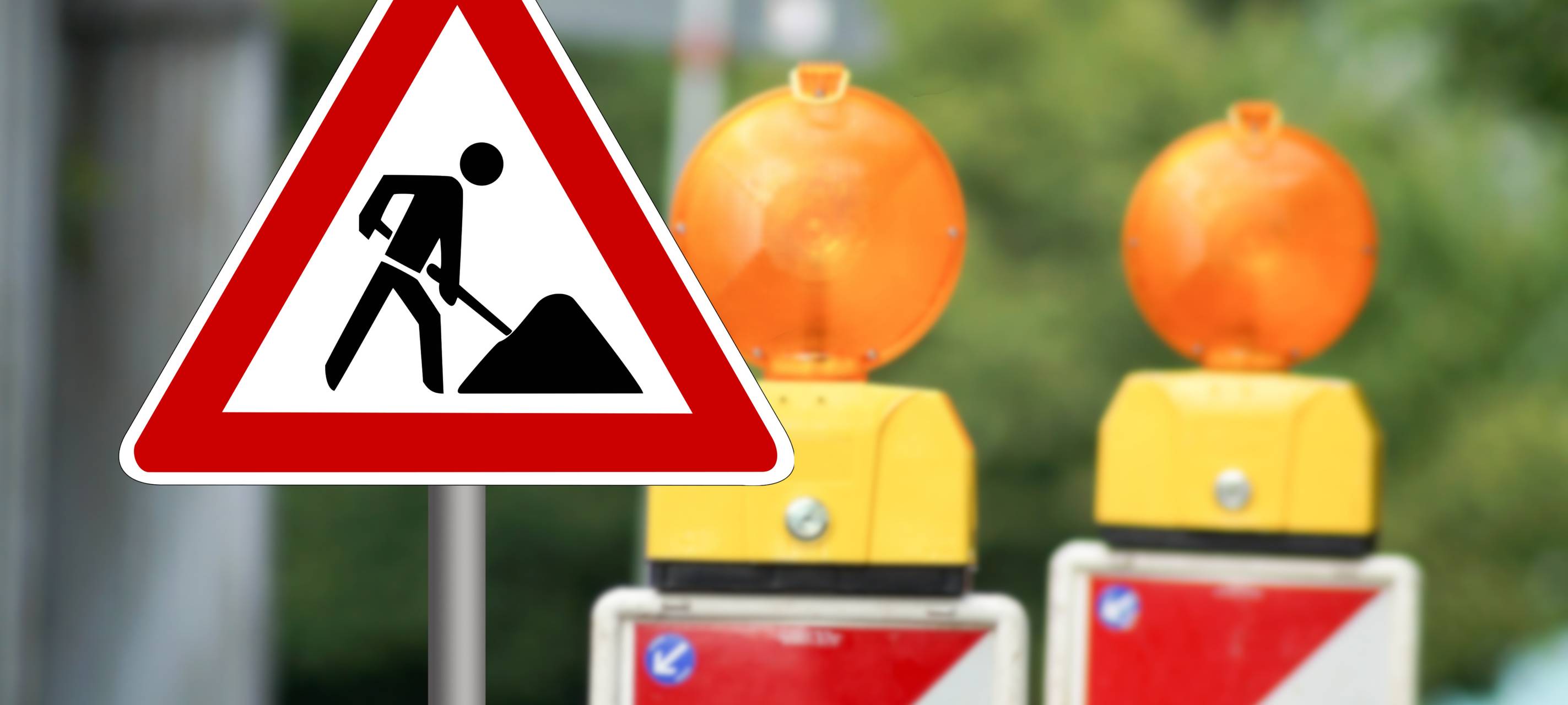 Ein Baustellenschild weist auf eine Baustelle hin. Im Hintergrund stehen zwei Warnbaken.
