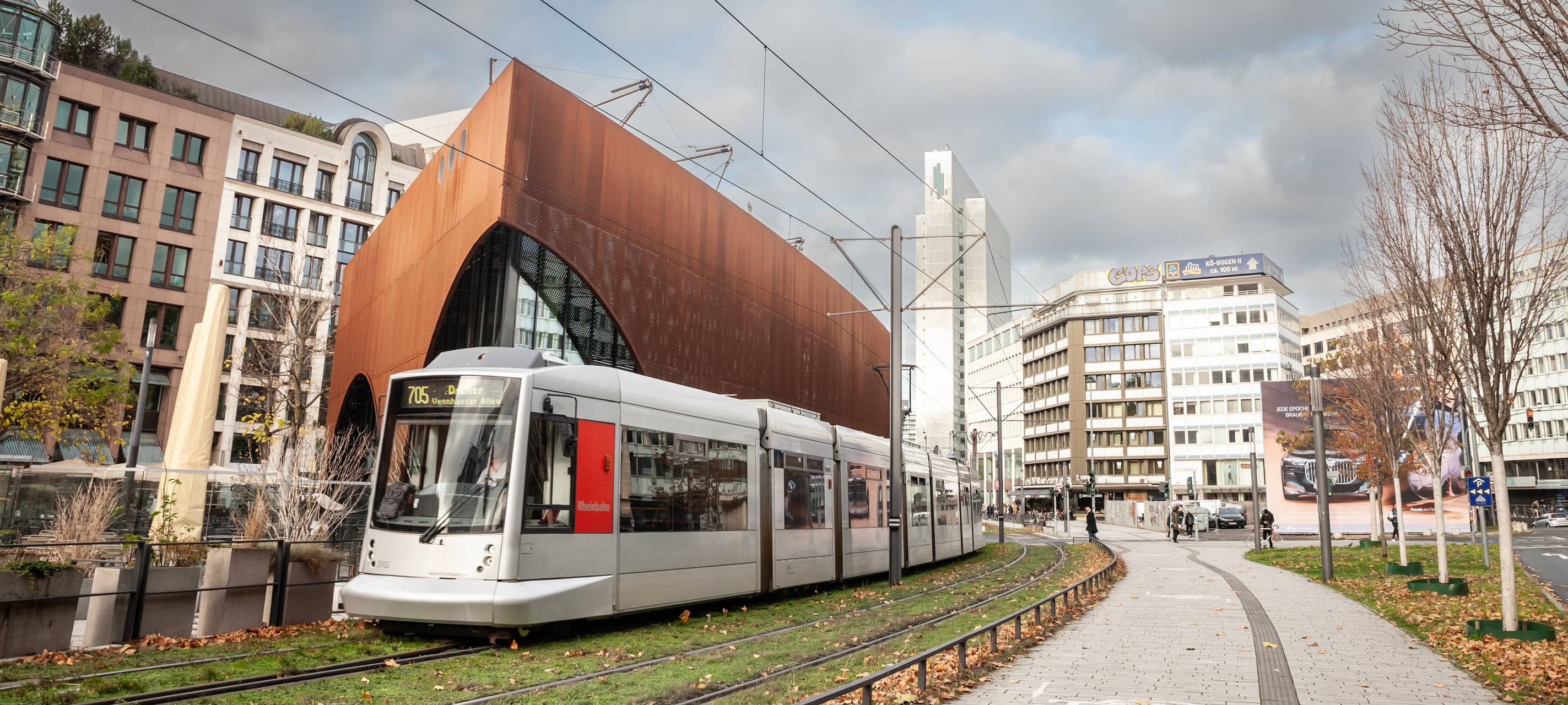 Eine Straßenbahn der Düsseldorfer Straßenbahn fährt durch das Stadtzentrum von Düsseldorf. Die Bahn wird von der Rheinbahn betrieben und ist Teil des öffentlichen Nahverkehrs. Im Hintergrund sind städtische Gebäude und Passanten zu sehen.