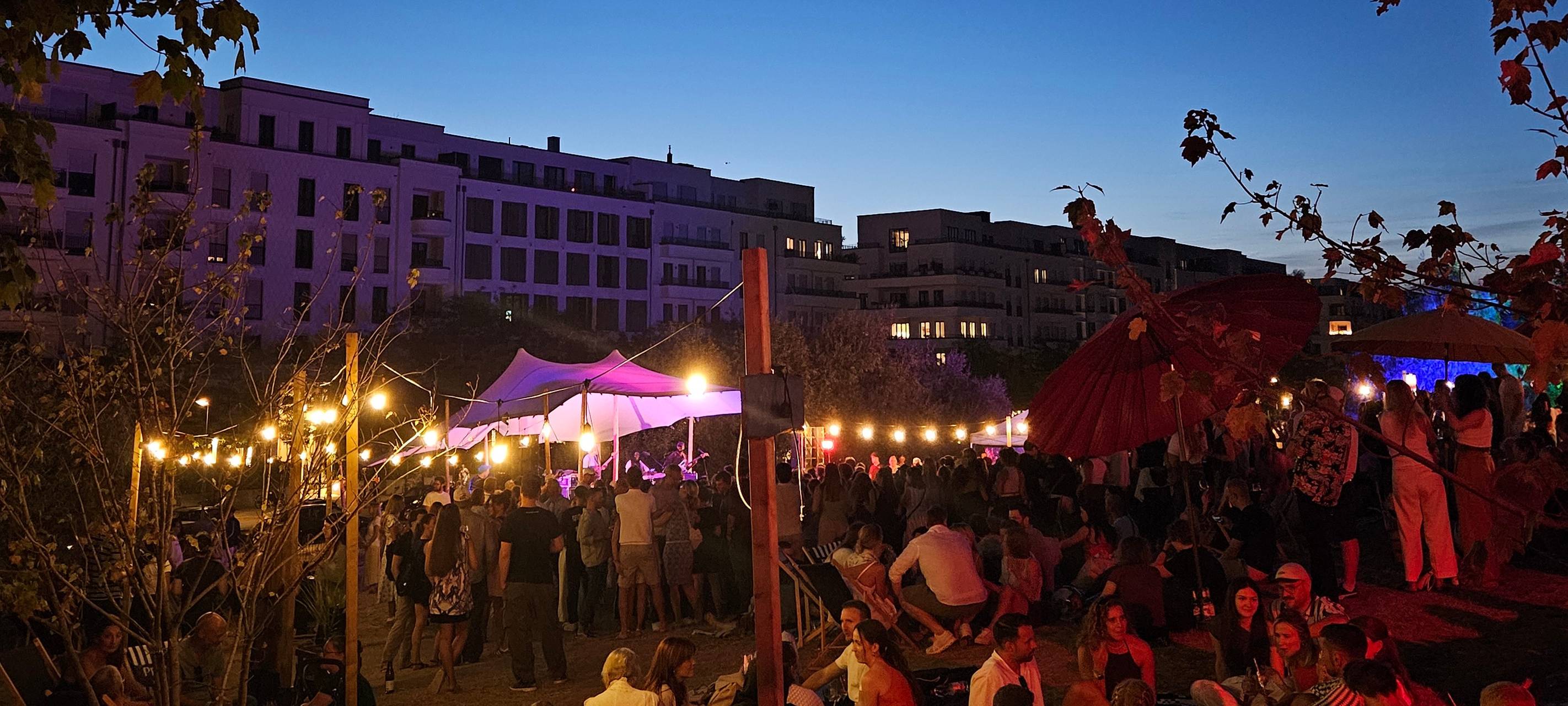 Das Liebe das Leben Festival bei Nacht. Vorne sitzen viele Menschen gemütlich zusammen. Im Hintergrund sind Wohnhäuser und der dunkele Nachthimmel zu sehen.