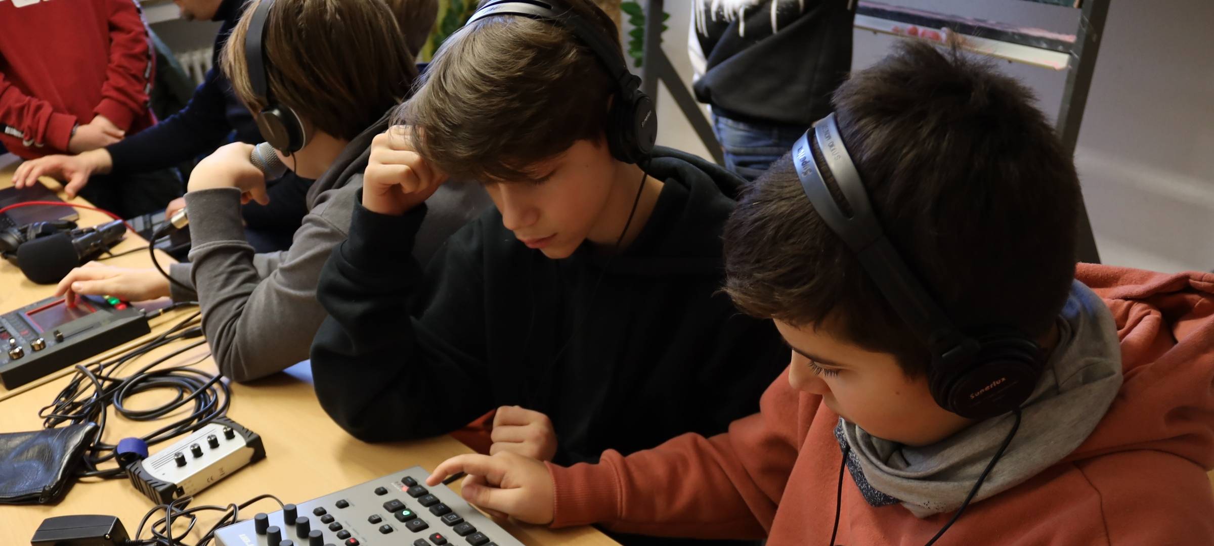 Mehrere Kinder sitzen vor einem Tisch und kleinen DJ-Pults. Die Kinder haben Kopfhörer auf und probieren das Pult aus.